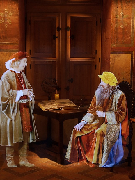 Two historical figures in period clothing at Clos Lucé, with a Mona Lisa replica nearby.