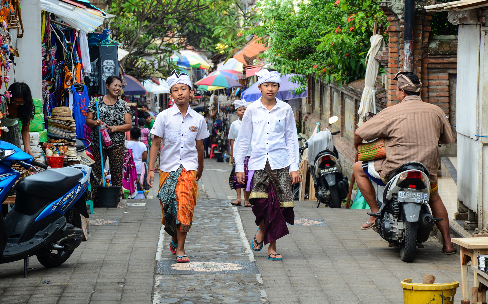 Boys in traditional attire walking through a bustling street market in Ubud, Bali.