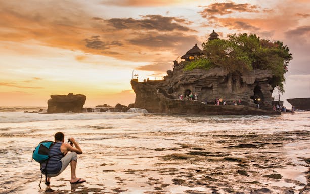 Tourist photographing Tanah Lot Temple at sunset, Bali, during a guided tour.