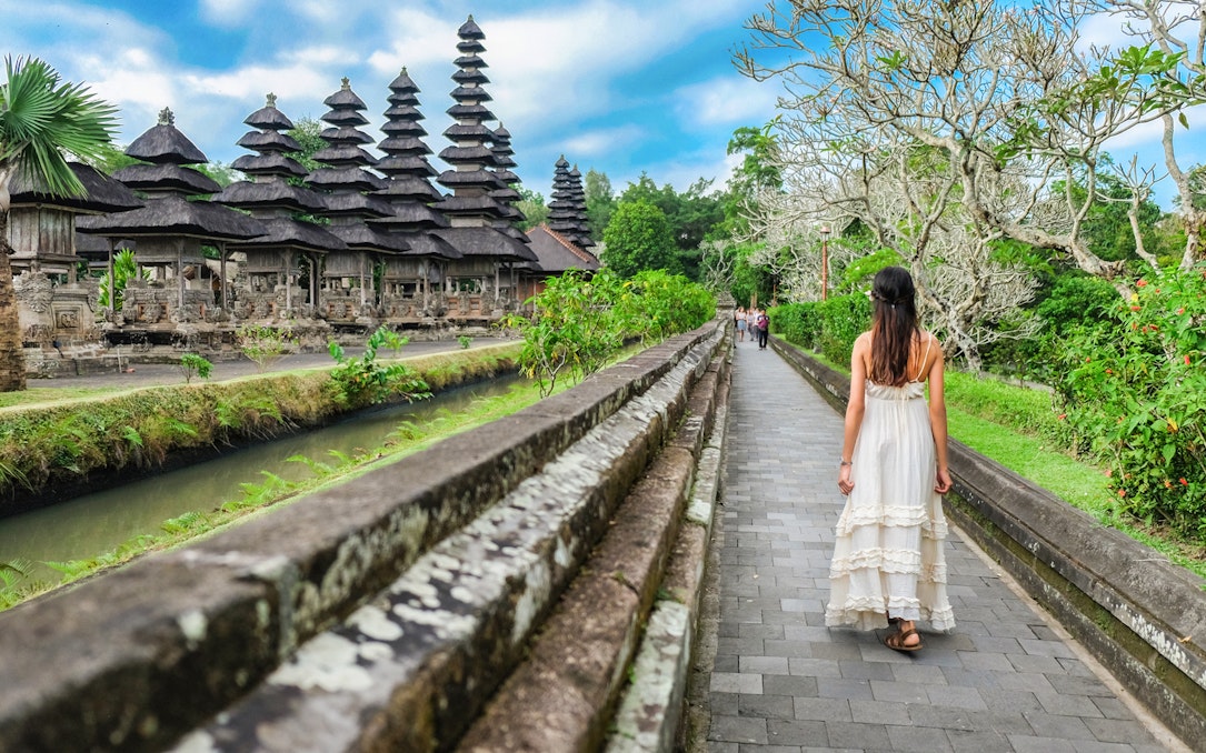 Woman walking along path towards Tanah Lot Temple, Bali, surrounded by lush greenery.