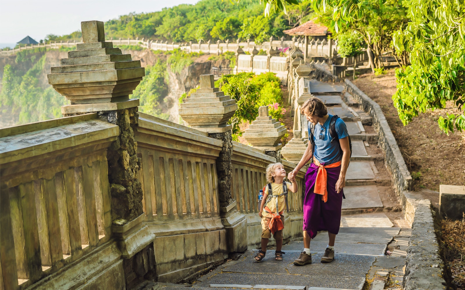 Father and child walking along Uluwatu Temple path in South Bali, surrounded by lush greenery.