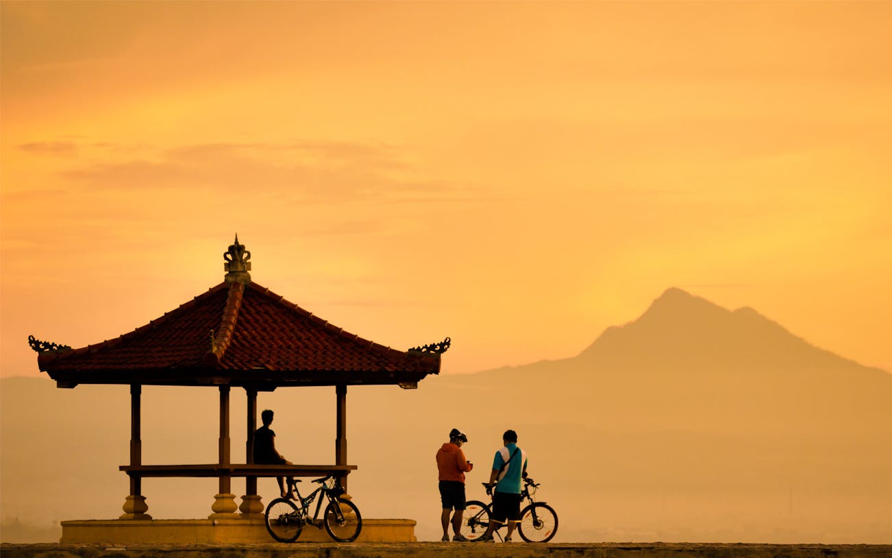 Cyclists at Sanur Beach gazebo during sunrise, Bali, with mountain view.