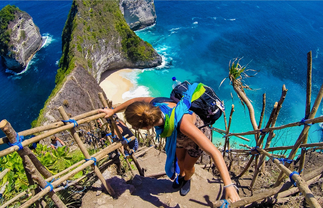 Hiker descending steep stairs to Kelingking Beach, West Nusa Penida, with ocean view.