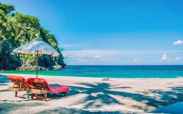Sun loungers and umbrella on Blue Lagoon Beach, Bali, with clear blue water and lush cliffs.