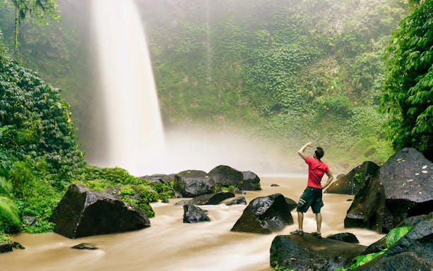 Person standing near Nungnung Waterfall surrounded by lush greenery, Bali.