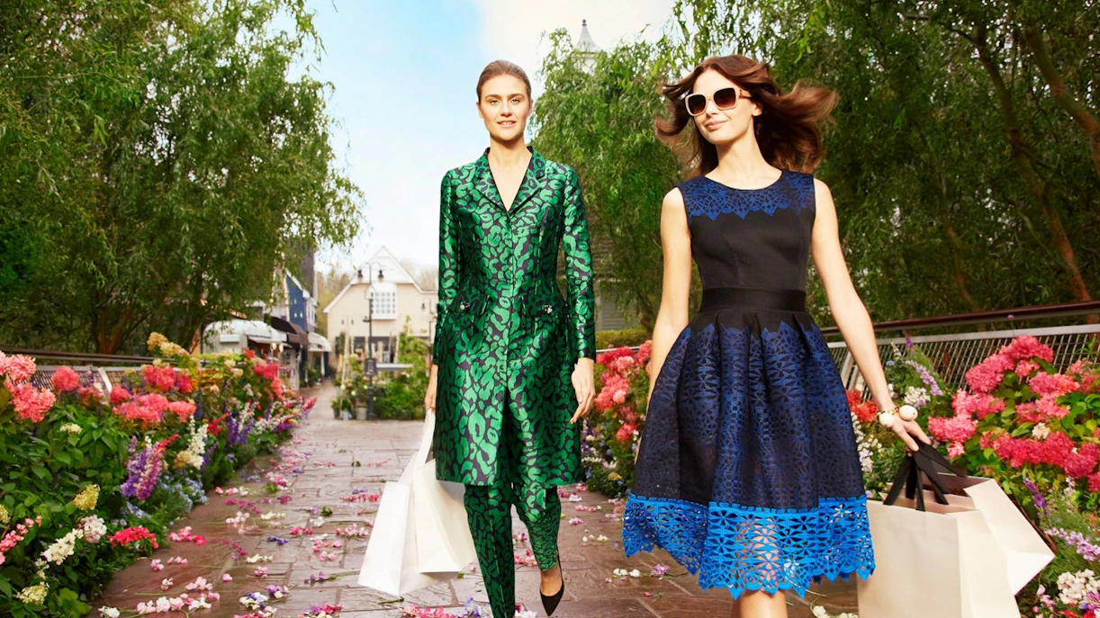 Shoppers walking through Bicester Village with shopping bags, surrounded by flowers.