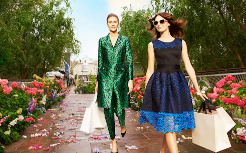 Shoppers walking through Bicester Village with shopping bags, surrounded by flowers.