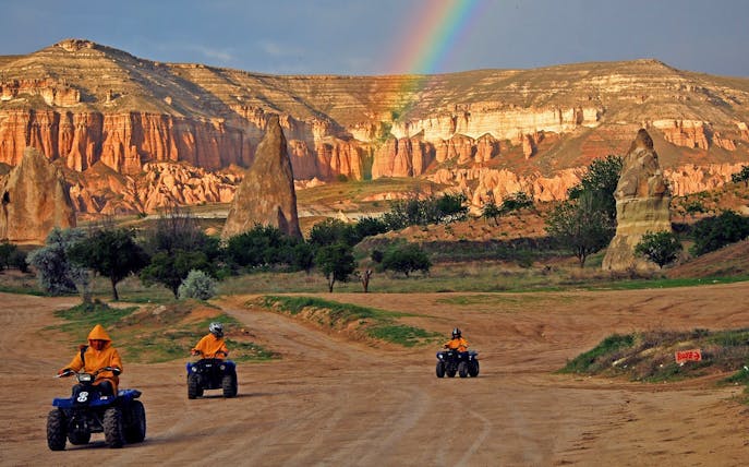 Quad bikes on a dirt path in Cappadocia with rock formations and a rainbow in the background.