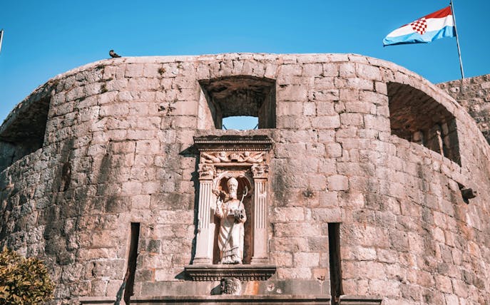Stone fortress wall with a statue and Croatian flag in Dubrovnik Old Town.