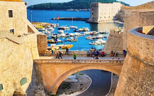 Dubrovnik city walls overlooking the harbor with boats and tourists on a bridge.