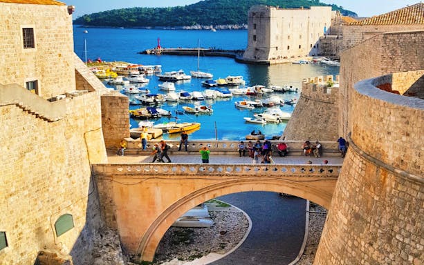 Dubrovnik city walls overlooking the harbor with boats and tourists on a bridge.