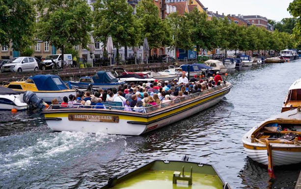 Boat tour on Copenhagen canal with tourists, passing colorful buildings and moored boats.