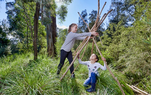 Children building a stick shelter at Healesville Sanctuary, surrounded by lush greenery.