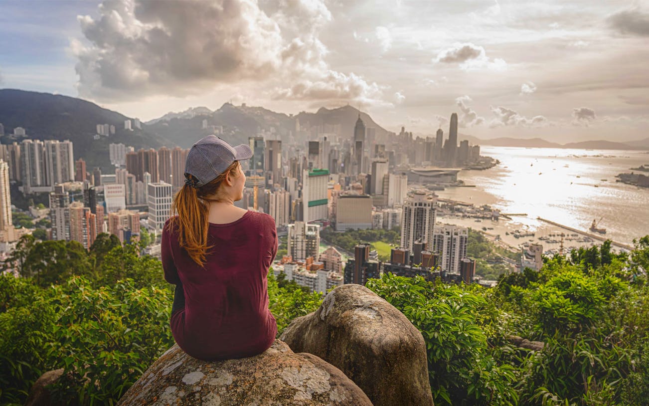 Person sitting on rock overlooking Hong Kong skyline from Victoria Peak during evening hike.