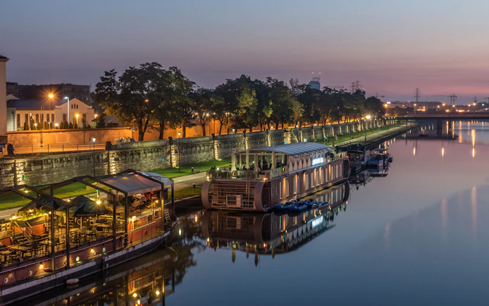 Krakow riverside at dusk with illuminated boats and tree-lined promenade.