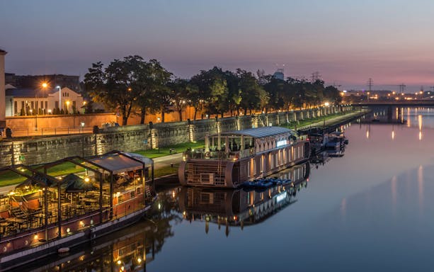 Krakow riverside at dusk with illuminated boats and tree-lined promenade.