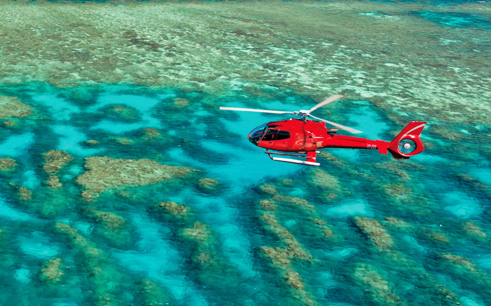 Red helicopter flying over the Great Barrier Reef's turquoise waters.
