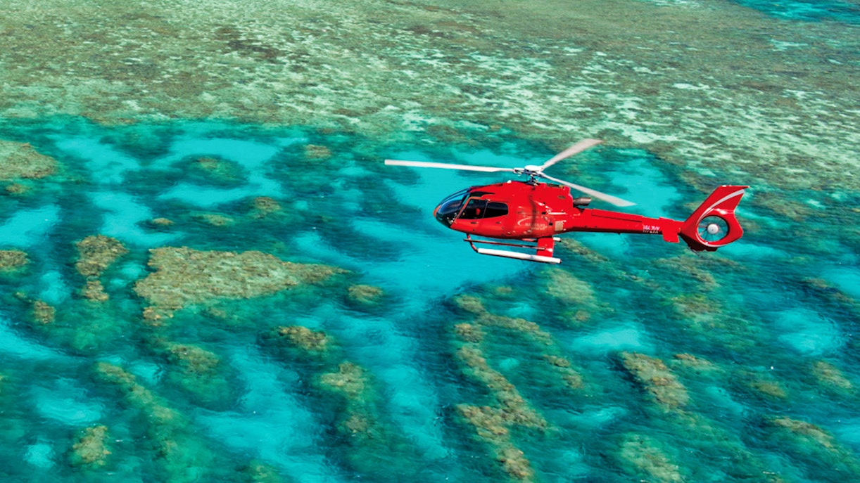 Red helicopter flying over the Great Barrier Reef's turquoise waters.