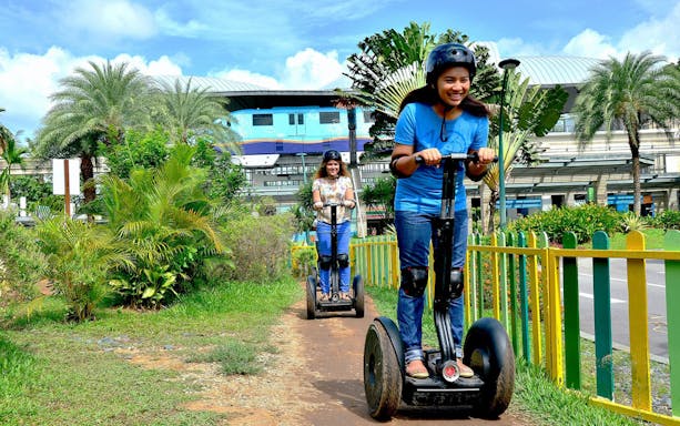Visitors riding Segways on a path in Sentosa, Singapore, with lush greenery and a monorail station in the background.