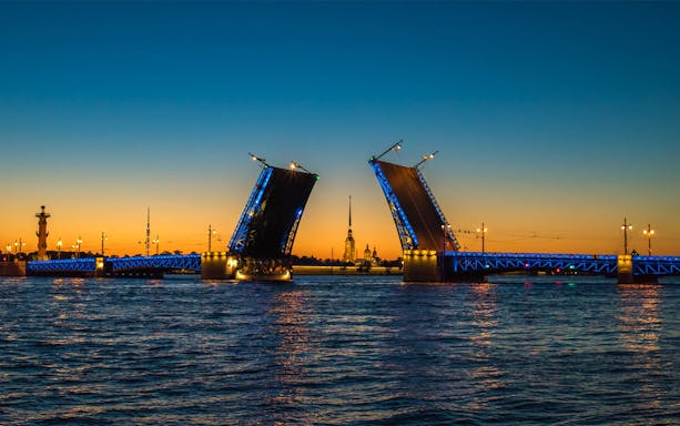 Drawbridge opening over river at sunset, with illuminated cityscape in the background.