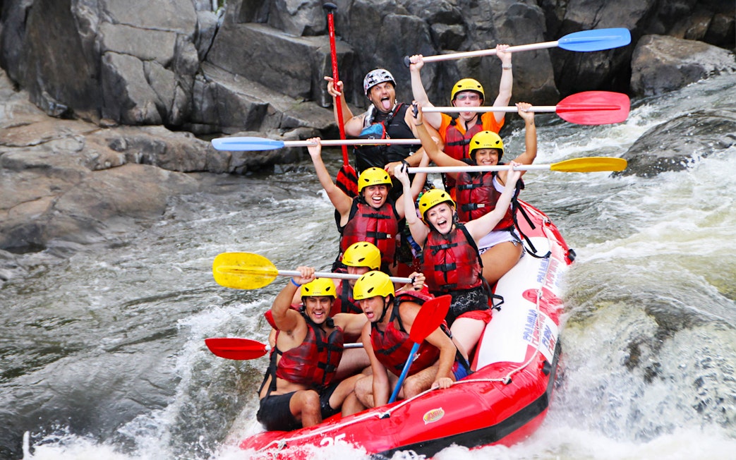 Group rafting on Barron River, navigating rapids with paddles raised, wearing helmets and life vests.