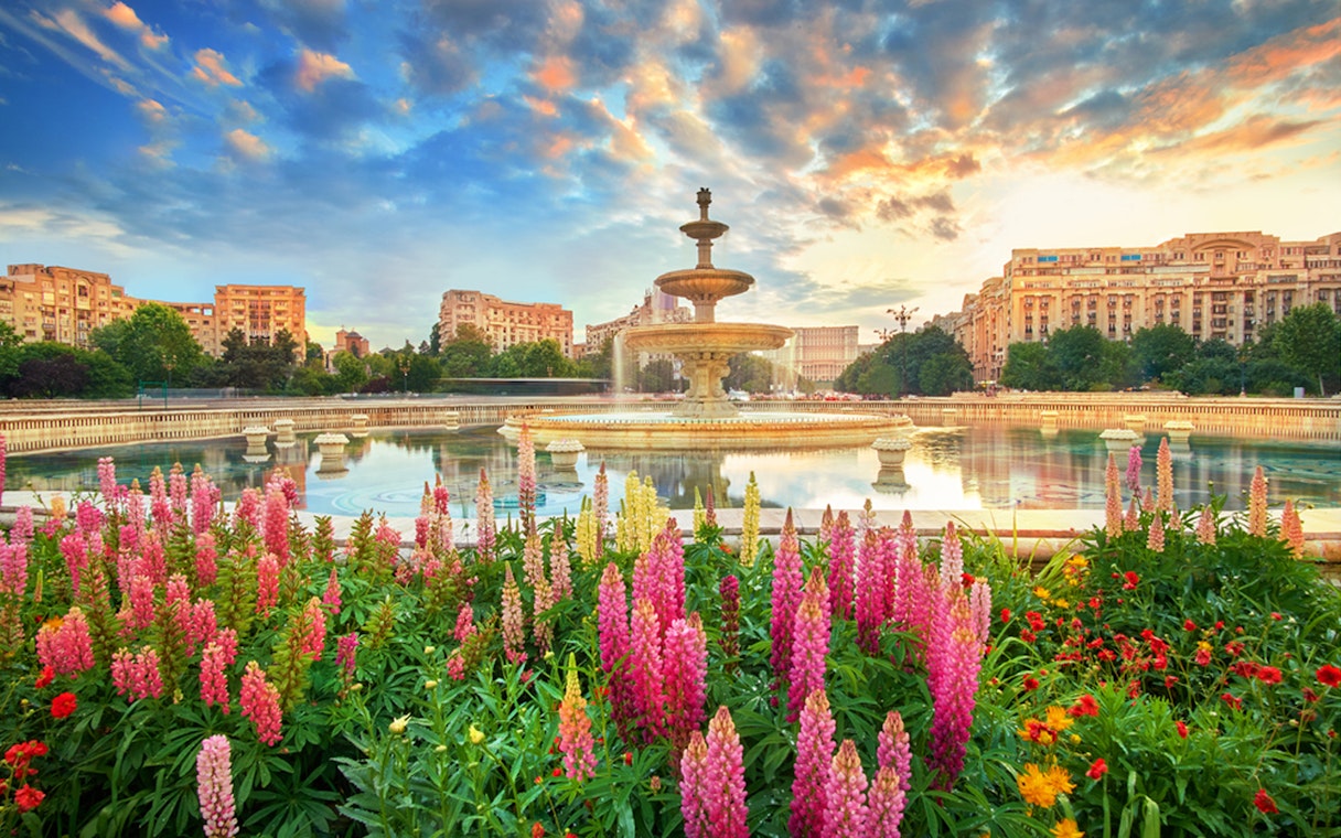 Fountain in Bucharest surrounded by colorful flowers at sunset, part of a private sightseeing tour.