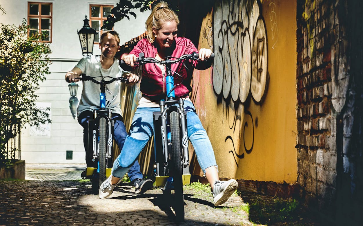 Two people riding e-scooters through a graffiti-lined alley in Prague.