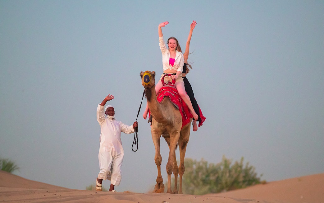 Camel ride in Dubai desert with guide and tourist waving.