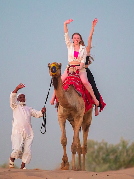 Camel ride in Dubai desert with guide and tourist waving.