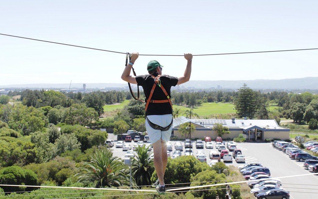 Person on high ropes course at West Beach Adventure, overlooking greenery and parking area.