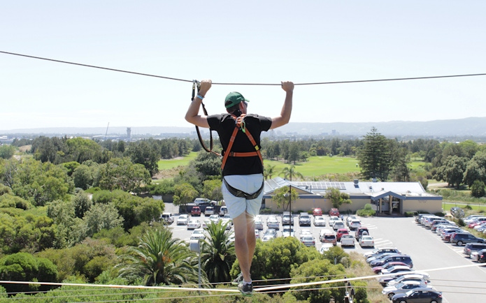 Person on high ropes course at West Beach Adventure, overlooking greenery and parking area.
