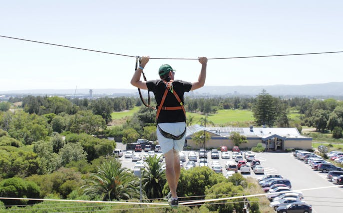 Person on high ropes course at West Beach Adventure, overlooking greenery and parking area.