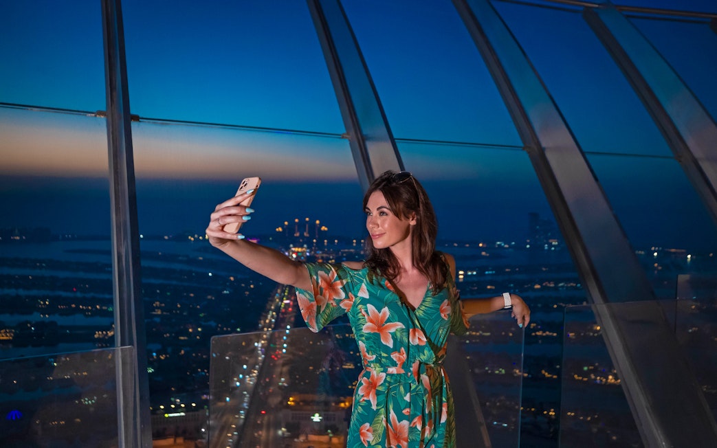 Person taking a selfie at The View At The Palm, Dubai, with city lights in the background.