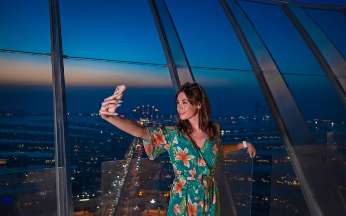 Person taking a selfie at The View At The Palm, Dubai, with city lights in the background.