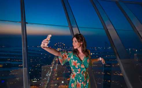 Person taking a selfie at The View At The Palm, Dubai, with city lights in the background.