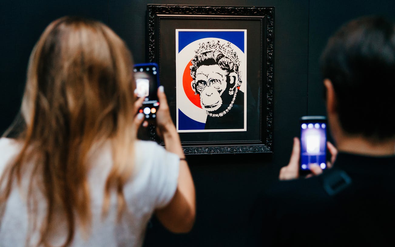 Visitors photographing Banksy artwork of a monkey with a crown at an exhibition.