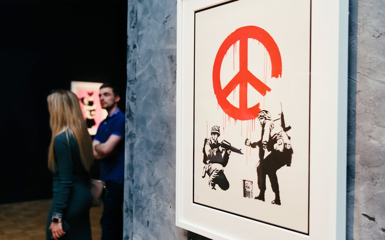 Banksy artwork with red peace symbol at exhibition, visitors observing.