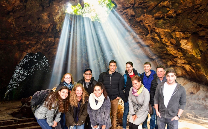 Group exploring Waitomo Glowworm Caves with light streaming through cave opening, Auckland tour.