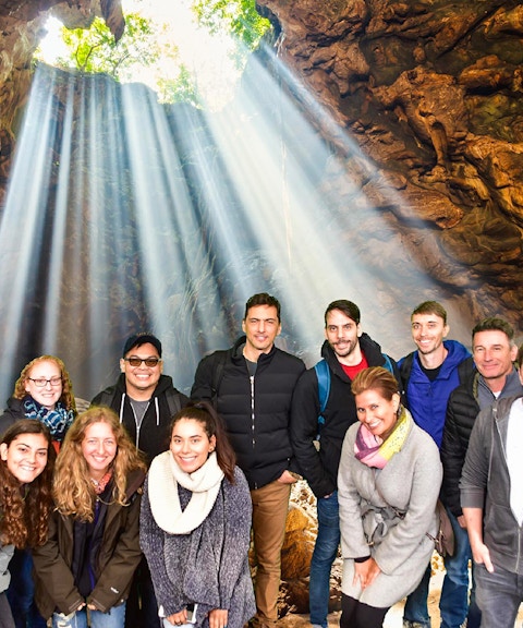 Group exploring Waitomo Glowworm Caves with light streaming through cave opening, Auckland tour.