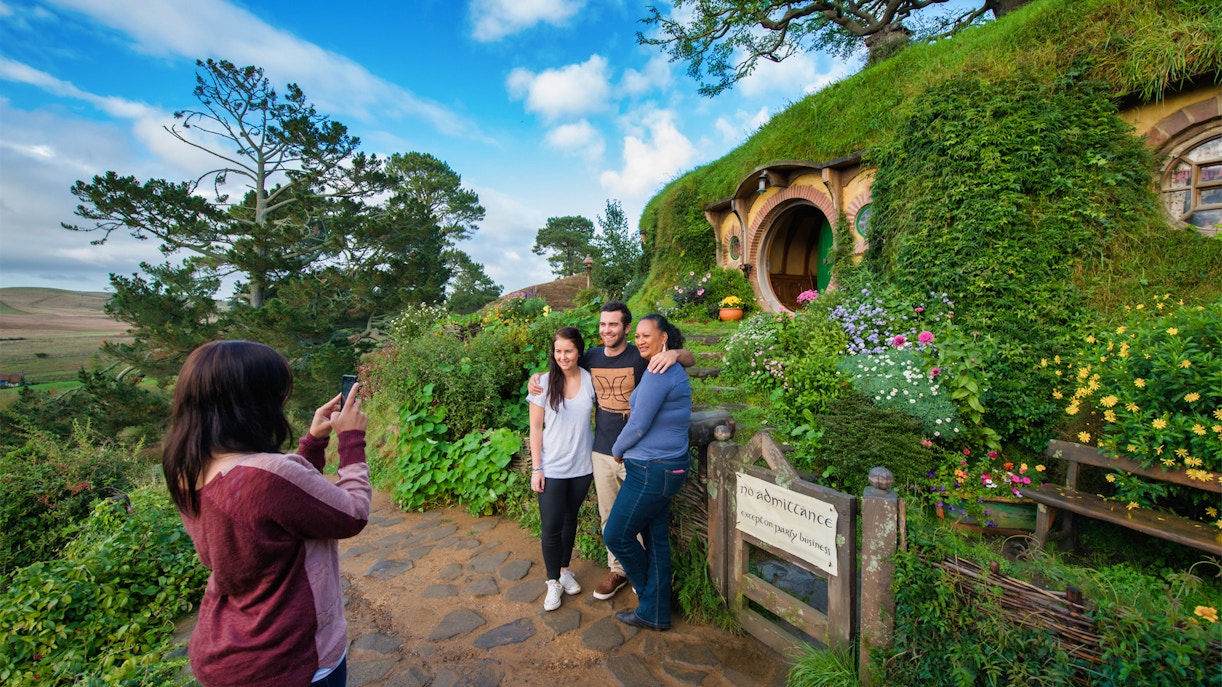Visitors posing at Hobbiton Movie Set, New Zealand, with iconic hobbit house in background.