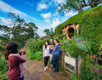 Visitors posing at Hobbiton Movie Set, New Zealand, with iconic hobbit house in background.