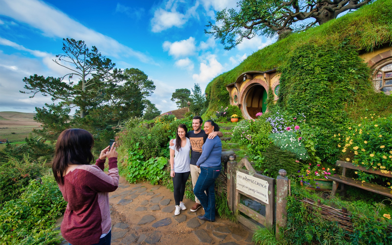 Visitors posing at Hobbiton Movie Set, New Zealand, with iconic hobbit house in background.