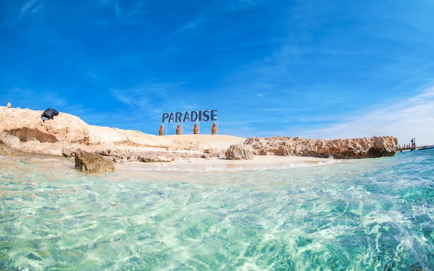 Giftun Island beach with clear water and "Paradise" sign, Egypt snorkeling tour.