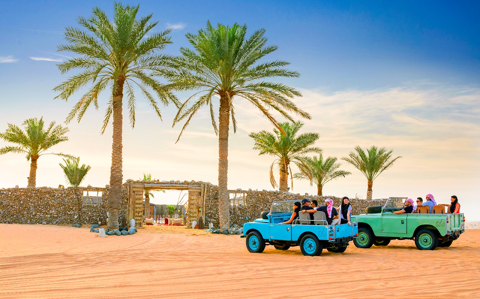 Vintage jeeps with tourists in a desert landscape, palm trees, and stone gate, Premium Heritage Desert Safari.