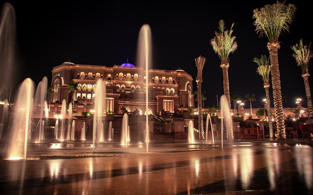 Fountains and palm trees in front of illuminated Emirates Palace, Abu Dhabi at night.