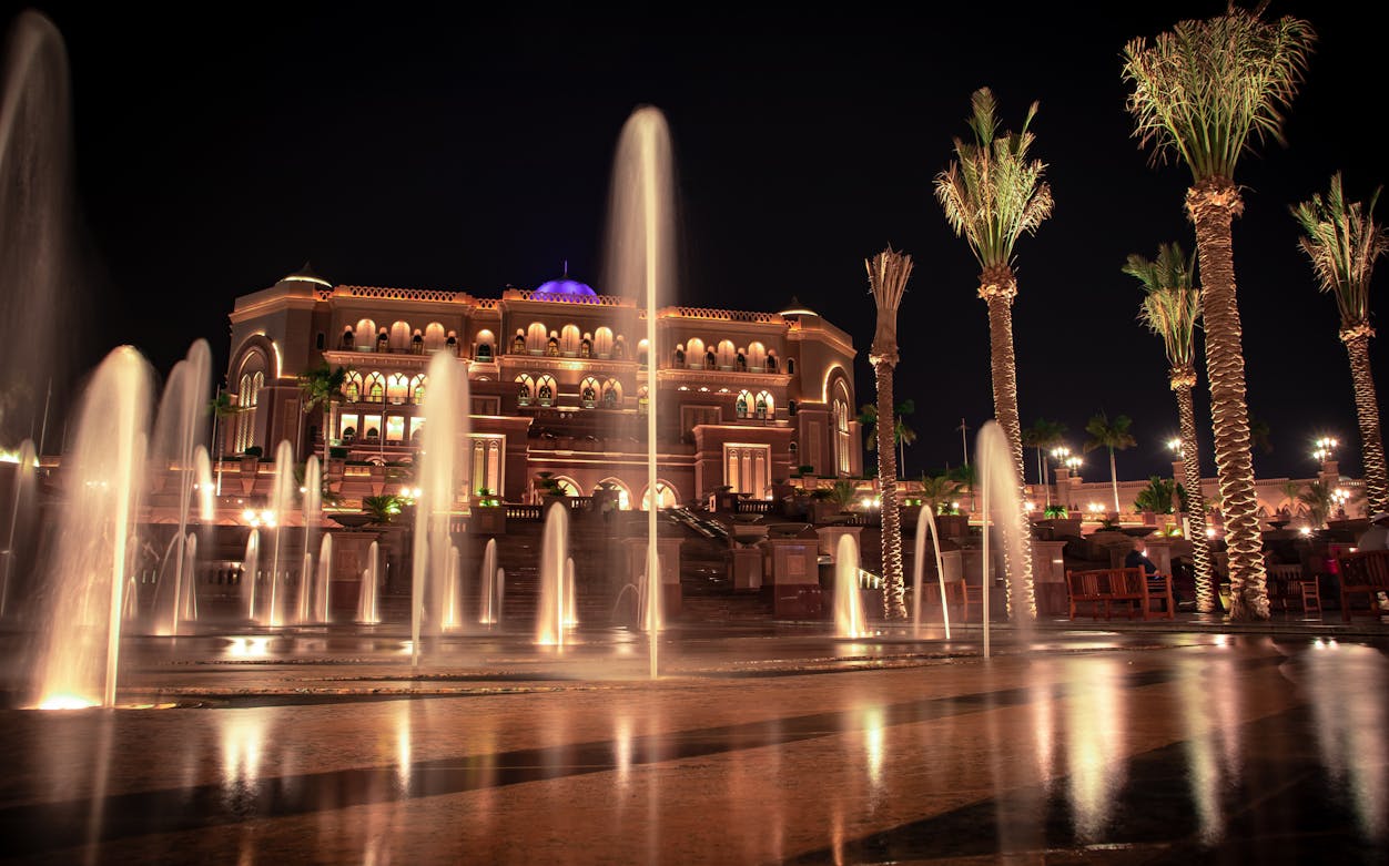 Fountains and palm trees in front of illuminated Emirates Palace, Abu Dhabi at night.