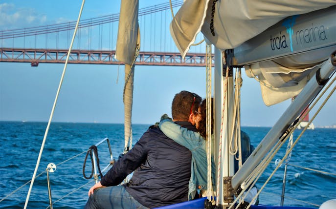 Couple embracing on a sailboat with Lisbon's 25 de Abril Bridge in the background.