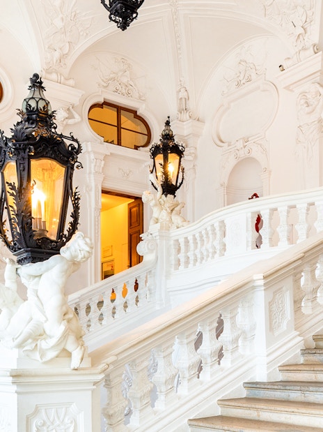 Upper Belvedere Palace interior staircase with ornate lanterns and sculptures.