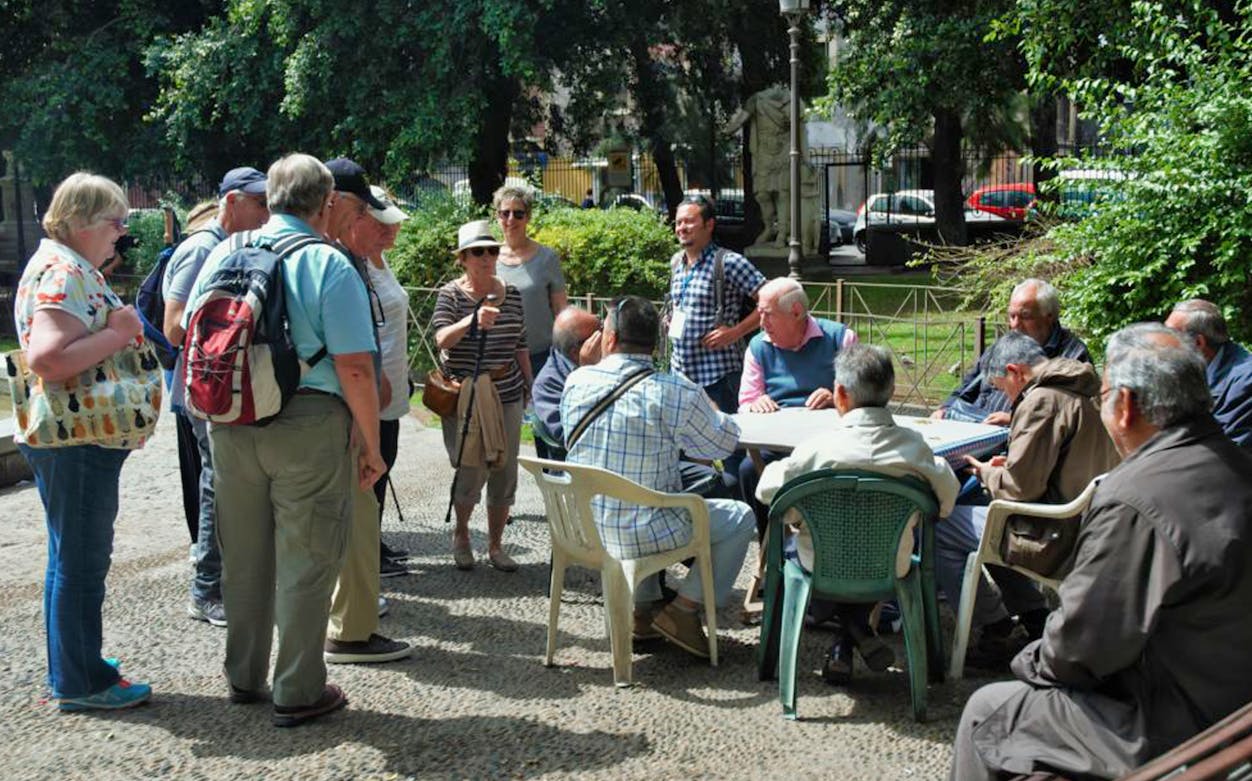 Group of tourists observing locals playing cards in a Catania park during a street food tour.