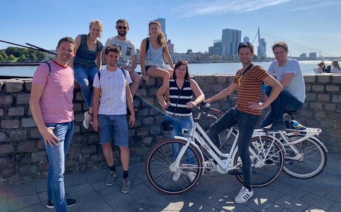Group of people on a bike tour in Rotterdam with city skyline and Erasmus Bridge in the background.